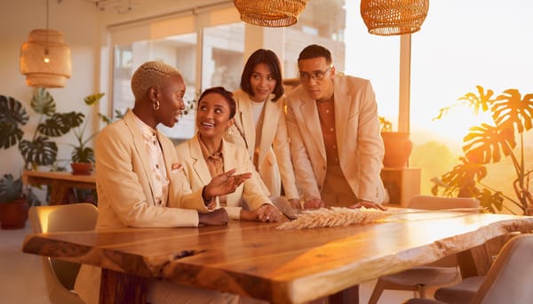 Four colleagues gathered at a long wooden table, looking down at plans spread across it, warm late sun behind them.