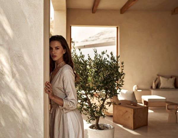 A woman in linen standing at a doorway beside a young olive tree, warm light in the room behind her.